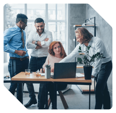 Group of four professionals collaborating around a laptop in a bright, modern office space with natural light and plants